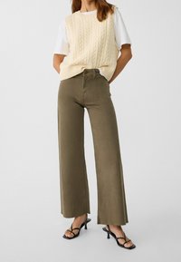 Beige kabelgebreide sweatervest boven een wit T-shirt, gecombineerd met olijfgroene wijde broekspijp en zwarte sandalen met bandjes, met rafelige mouwen.