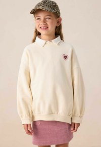 Crème-kleurige sweatshirt met ballonmouwen, voorzien van een roze hart-applicatie met een zwarte middenstuk. Gepaard met een roze corduroy rok.