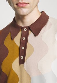 Muž oblečený v polo tričku s vlnitým vzorem v hnědé, béžové a hořčicové barvě se čtyřmi béžovými knoflíky a hnědým límcem, zobrazený od brady k hrudi.