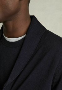 Sudadera de un oscuro azul marino con cuello chal, que presenta una textura acanalada y está superpuesta a una camisa gris claro por debajo. Vista de cerca del escote.