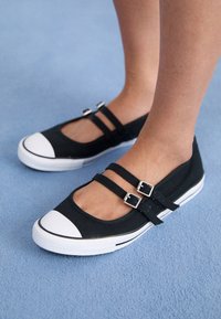 Pies de niños que llevan deportivas de lona negras tipo Mary Jane con puntas de goma blanca y doble correa en un suelo de moqueta azul.