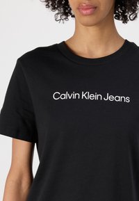 Melnas kokvilnas T-krekls ar īsām piedurknēm un apaļu kakla izgriezumu, ar baltu tekstu "Calvin Klein Jeans" priekšpusē.
