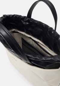 Tote bag crème avec poignées en cuir noir et haut à cordon, montrant l'intérieur en tissu beige avec poches cousues.