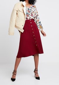 Een aansluitende bordeauxrode rok met een knoopsluiting aan de voorkant, gecombineerd met een bloemenblouse en een crèmekleurige spijkerjack. Zwarte sandalen met bandjes maken de look af.