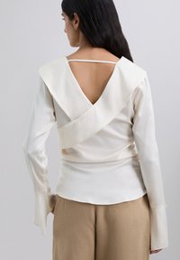 Blusa blanca roto con un diseño de espalda en V drapeada, mangas largas y puños acampanados, hecha de una tela suave y ligera.