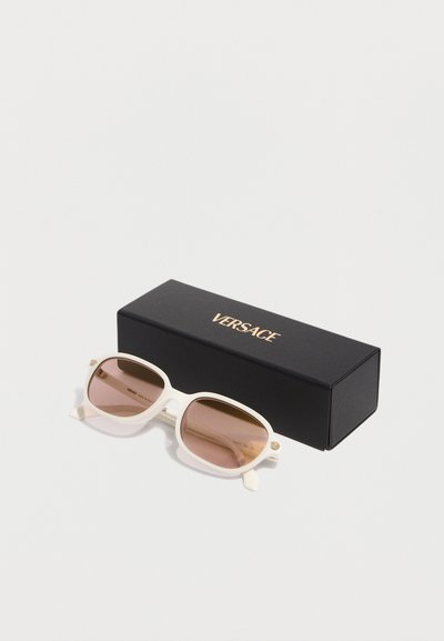 Γυαλιά ηλίου Versace με λευκό σκελετό και καφέ φακούς με βαθμιαία απόχρωση, τοποθετημένα δίπλα σε μια μαύρη συσκευασία με το λογότυπο Versace σε λευκό φόντο.