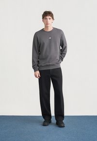 Γκρι φούτερ με στρογγυλή λαιμόκοψη και μικρό λογότυπο στο στήθος, συνδυασμένο με μαύρο παντελόνι φαρδύ και μαύρα παπούτσια. Απλό φόντο.