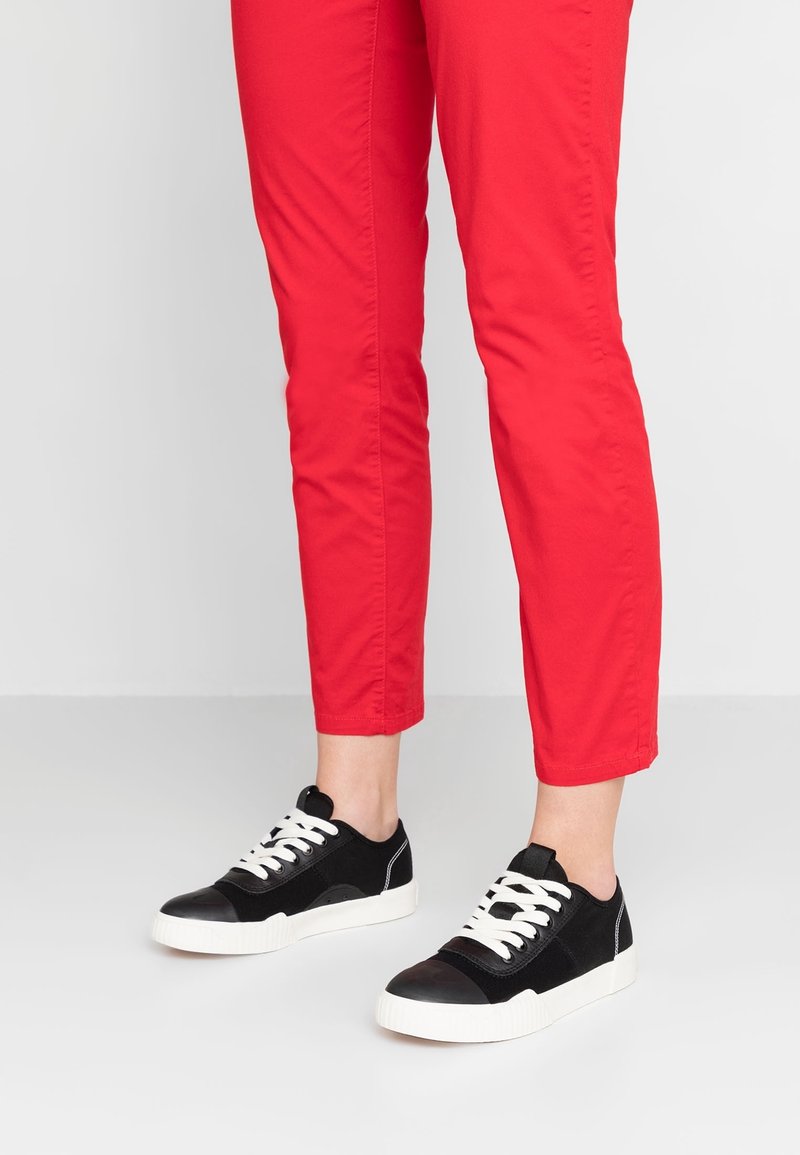 Personne portant un pantalon rouge et des baskets basses noires avec des lacets blancs, se tenant sur un sol blanc uni devant un fond blanc.