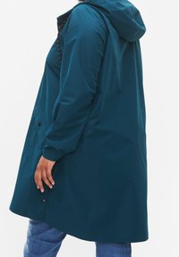 Teal vattentät jacka med huva, långa ärmar och sidofickor. Slät yta med knappdekorationer och justerbara dragsnören vid kragen.