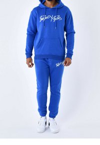 Set di felpa blu con cappuccio e pantaloni jogging in cotone, con tasca frontale e logo in corsivo bianco. Completato con sneaker bianche.
