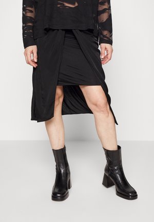 Zwarte lange mouwen top met doorschijnende, versleten accenten, gelaagd over een zwarte wikkelrok. Zwarte leren enkellaarsjes met een ritsluiting.