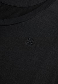 Close-up van zwarte crewneck stof met subtiele gestructureerde weving en een klein, geborduurd donker logo in het midden.