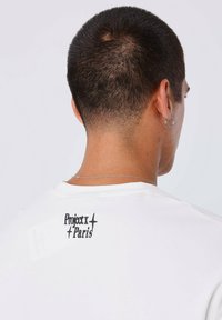 Camiseta blanca con un logotipo de texto gráfico negro "Project x Paris" en la parte superior de la espalda, con una textura de tela suave y lisa.