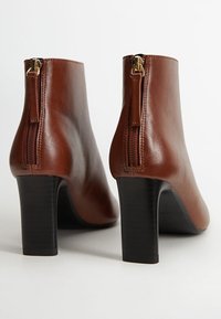 Bottines en cuir marron à texture lisse, avec un talon bloc haut et des fermetures éclair latérales visibles ornées de pièces en or.