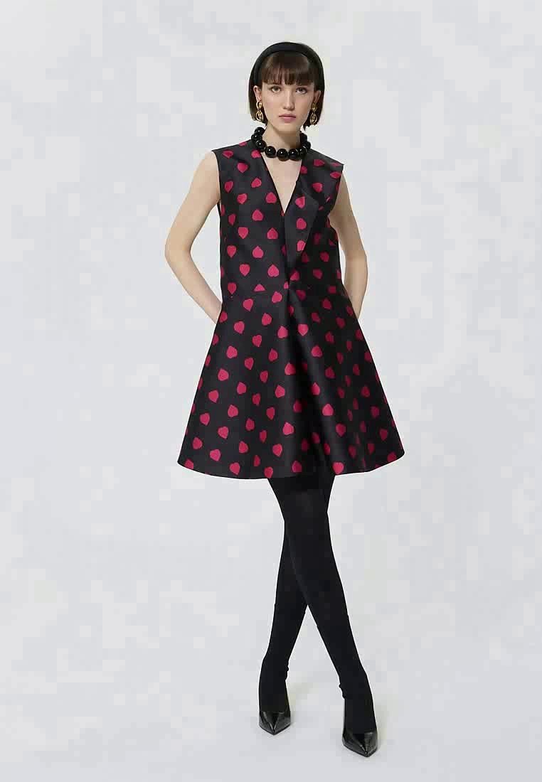 Femme portant une robe noire sans manches avec des pois rouges, des collants noirs, des talons noirs, un collier de perles noires et des boucles d'oreilles en or, se tenant avec les mains dans les poches.