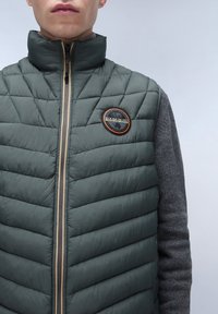 Padded grüner Weste mit hohem Kragen, gestepptem Muster und einem kreisförmigen Logo auf der Brust; verfügt über einen beigen Reißverschluss an einem grauen Ärmel.