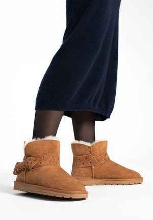 Botas de tobillo de ante en color marrón claro con un puño doblado, forro de piel sintética blanca y adornos de tachuelas decorativas. Suela plana y diseño de punta redonda.