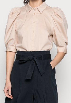 Donna che indossa una camicia button-up con maniche a palloncino beige chiaro, infilata in pantaloni scuri a vita alta, con una cintura in tessuto annodata.