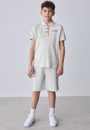 Fiú áll világosszürke galléros pólóban és hozzáillő rövidnadrágban, fehér tornacipőt és zoknit viselve, egyszínű világos háttér előtt.