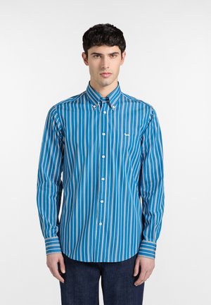 Junger Mann trägt ein blaues, langärmliges Hemd mit vertikalen weißen Streifen, steht vor einem schlichten weißen Hintergrund.