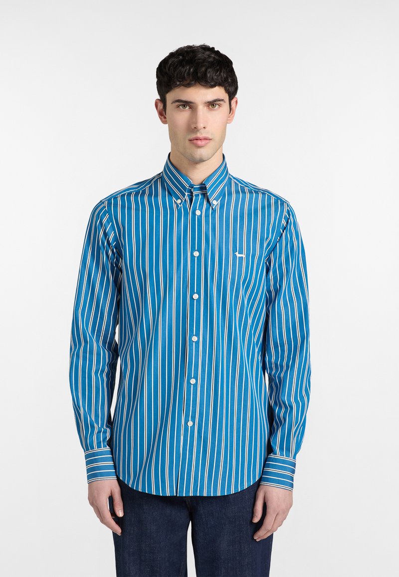 Jeune homme portant une chemise bleue à manches longues avec des rayures blanches verticales, debout devant un fond blanc uni.