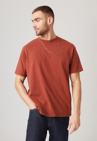 Camiseta de algodón color óxido con cuello redondo y mangas cortas. Presenta un pequeño logo en relieve en el pecho. Llevada con jeans de mezclilla oscura.