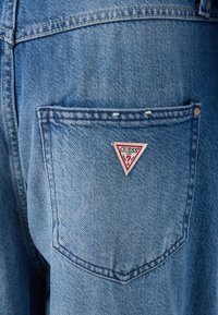 Jean en denim avec une poche arrière ornée d'un patch logo triangulaire "GUESS" en rouge et blanc, affichant un tissu bleu texturé et délavé.