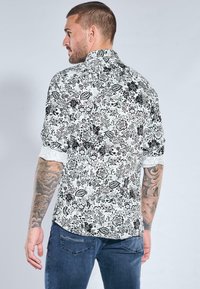 Schwarz-weiß gemustertes Kurzarmhemd mit floralen Motiven und tailliertem Schnitt. Verfügt über eine kontrastierende weiße Manschette und sichtbare Tätowierungen an den Armen.