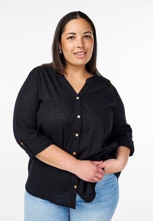 Camicia nera button-up con scollatura a V e maniche arrotolate, realizzata in un tessuto testurizzato. Bottoni in stile legno aggiungono un tocco di dettaglio. Indossata sopra dei jeans azzurri chiari.