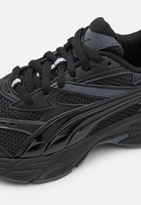 Black athletic shoe featuring mesh and synthetic upper, rounded toe, textured overlays, and flat laces. Sole has a wave pattern design.