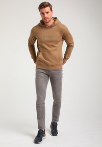 Bruine hoodie met een voorzak en logodetail, gecombineerd met slim-fit grijze jeans en marineblauwe sneakers. Vlakke, zachte textuur.