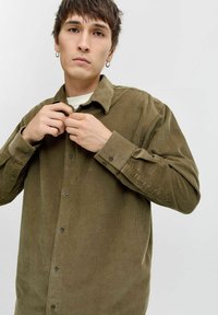 Giovane con capelli corti castani e orecchini a cerchio che allaccia una camicia di velluto a coste marrone sopra una maglietta bianca, contro uno sfondo neutro.