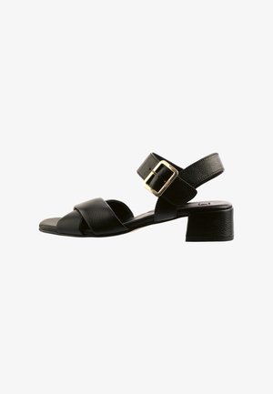 Zwarte leren sandalen met een lage blokhak, voorzien van gekruiste banden en een gespsluiting rond de enkel. Textuurafwerking.
