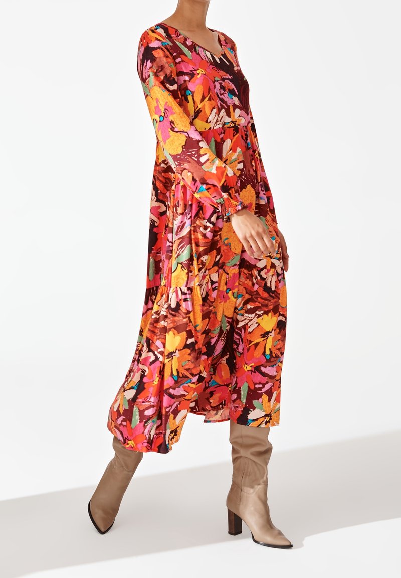 Robe longue à motifs floraux avec des couleurs vives de rose, orange et vert, dotée de longues manches et d'un décolleté en scoop, assortie à des bottes beiges arrivant au genou.