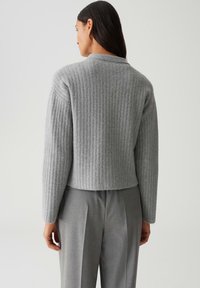 Femme aux longs cheveux bruns portant un pull en maille gris et un pantalon gris ajusté, vue de dos sur un fond uni.
