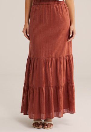 Fustă maxi în nuanțe ruginii, cu volum și dungi verticale subtile, purtată cu sandale deschise pe deget bej, pe un fundal luminos.