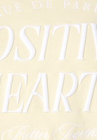 Textiel met een zachte, lichtgele stof met verhoogde witte letters, waaronder "POSITIEF" en "HART." De tekst is duidelijk gestikt met een gladde textuur.
