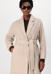 Cappotto in misto lana beige con cintura, rever a punta, tasche laterali e superficie testurizzata. Indossato sopra un maglione marrone scuro.