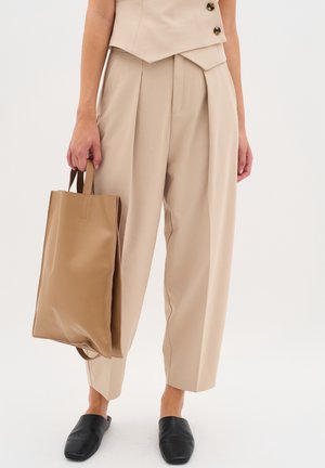 Person iført beige højtaljede plisserede bukser, der holder en tan læder tote taske og iført sorte slip-on sko på en lys baggrund.