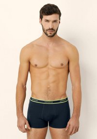 Boxeri scurți de culoare albastru închis cu o textură moale, având un tiv elastic de culoare verde neon inscripționat "yamamay." Design ajustat, fără modele vizibile.