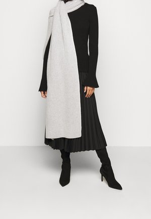 Femme portant une longue écharpe grise claire côtelée sur un pull noir côtelé, une jupe noire plissée et des bottes à talons noires, sur un fond uni.