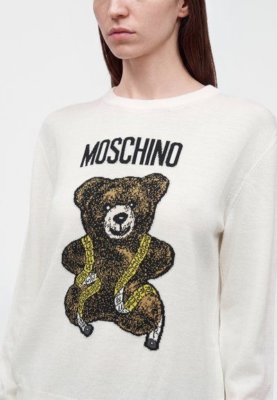 Personne portant un pull blanc avec un ours en peluche marron enveloppé dans un ruban à mesurer jaune avec le mot Moschino au-dessus.