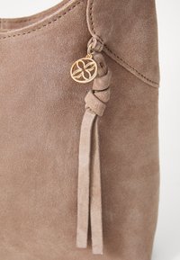 Sac à main en daim marron avec un gland noué et un accent en forme de fleur dorée. Texture douce avec des bords lisses et sans matériel visible.