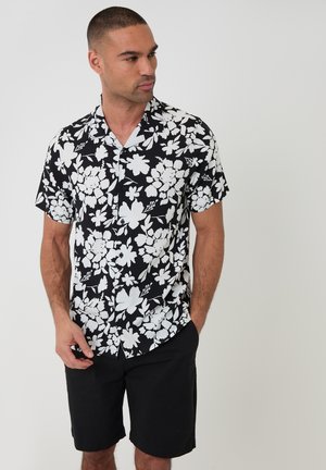 Mężczyzna ubrany w czarno-białą koszulę na krótki rękaw z kwiatowym wzorem i czarne szorty, stojący z częściowo włożonymi rękami do kieszeni.