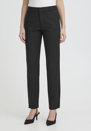 Kobieta ubrana w czarne, dopasowane spodnie o długości do kostek i czarne szpilki z zaostrzonymi czubkami na jednolitym tle.