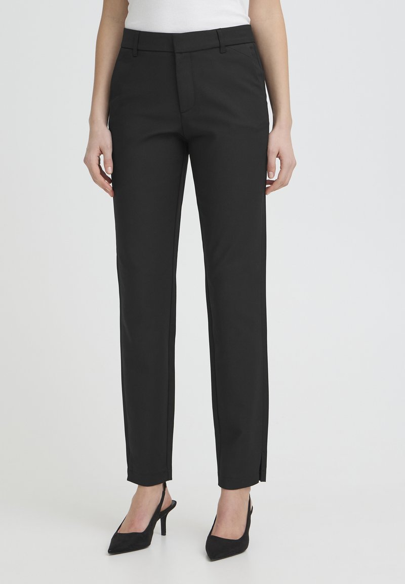 Femme portant un pantalon noir sur mesure à longueur cheville et des talons noirs à bout pointu sur un fond uni.