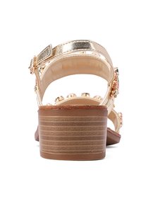 Sandal in metallo dorato con cinturino posteriore testurizzato in oro, suola piatta e gioielli decorativi sul cinturino. Presenta un tacco basso e scanalato.