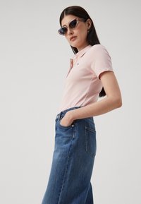 Svijetlopinkasta polo majica s malim logom, u kombinaciji s trapericama koje imaju visoki struk. Model nosi velike mačje naočale.