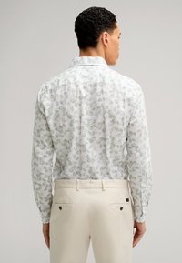 Langärmliges Hemd in Weiß mit grünem Blumenmuster, mit Button-Down-Kragen und Detail an den Manschetten; kombiniert mit hellbeigen Hosen.