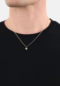 Collana in oro con un piccolo ciondolo a forma di cuore, indossata sopra una camicia nera, che mostra una finitura liscia e lucida.